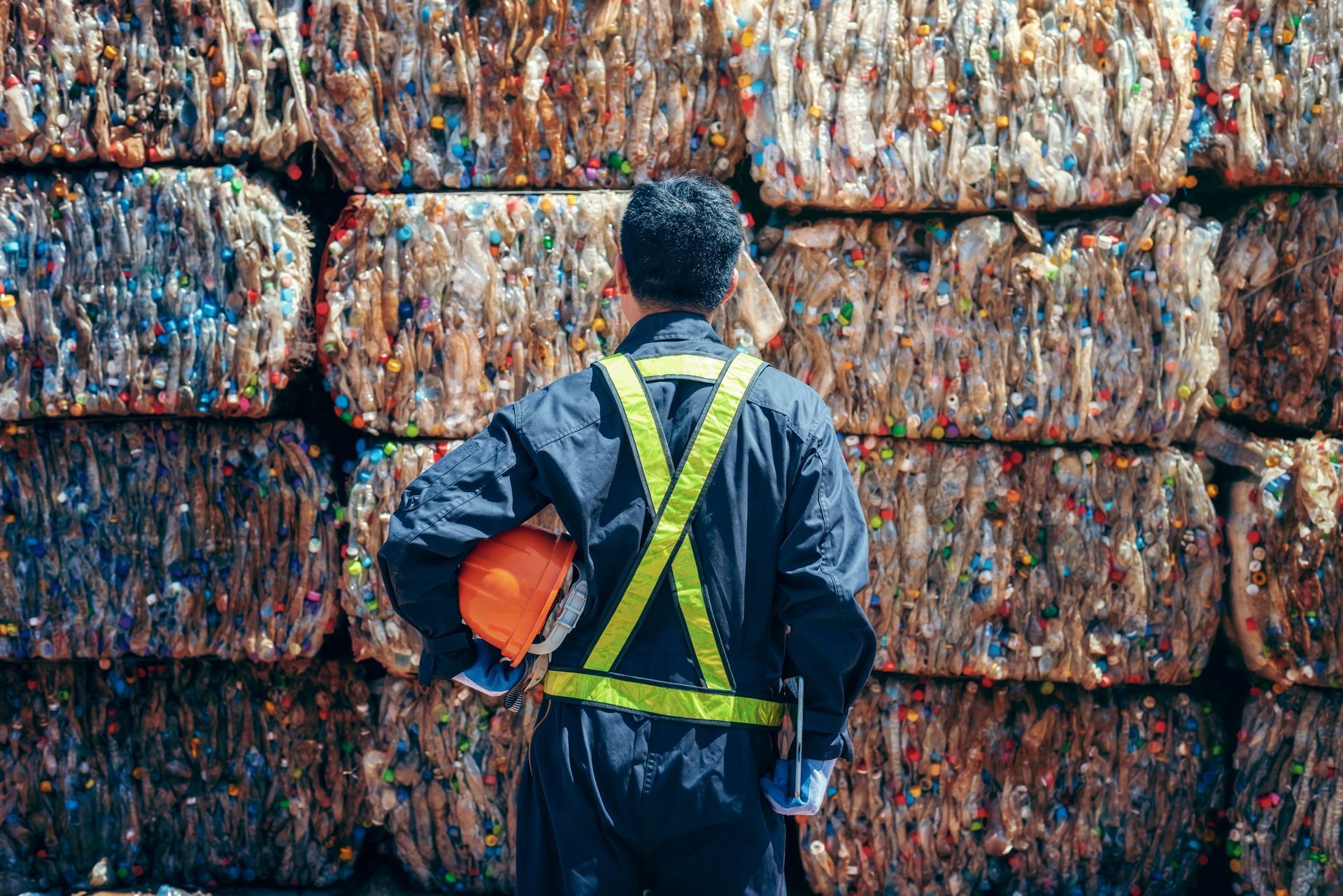 Man in safety vest holding a hard hat stands before stacked, compressed bales of recycled material. Man in safety vest holding a hard hat stands before stacked, compressed bales of recycled material.