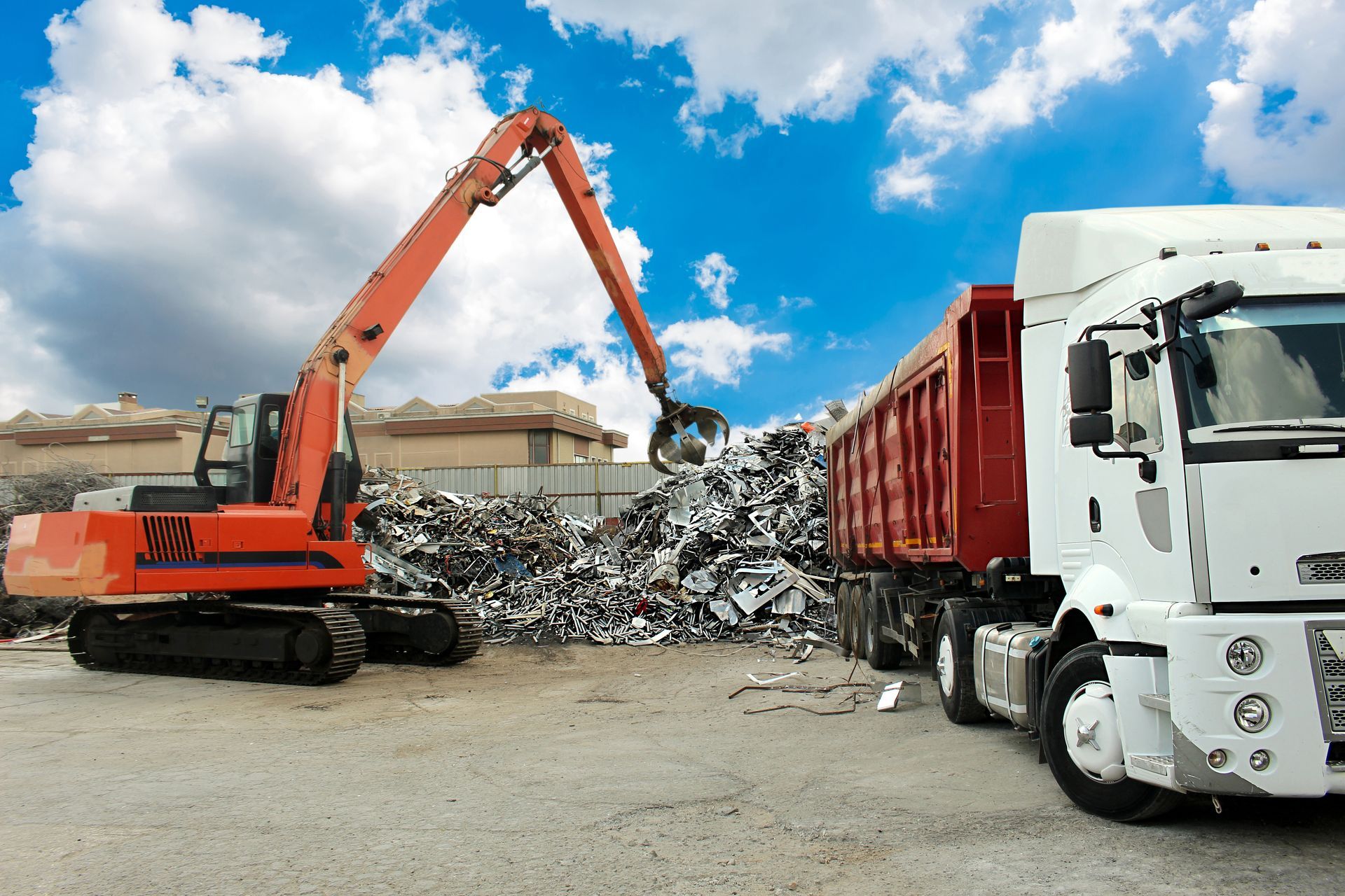 Orange excavator loading scrap metal into a red trailer truck at a recycling yard under a cloudy sky. Orange excavator loading scrap metal into a red trailer truck at a recycling yard under a cloudy sky.