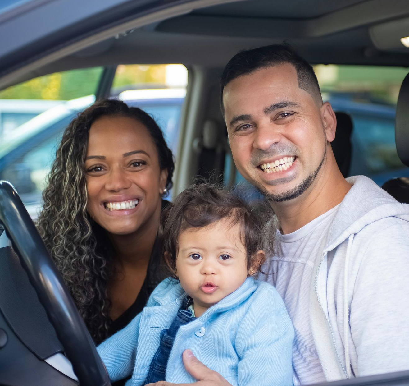 Family of three smiling in a car. Child in blue jacket held by adults.
