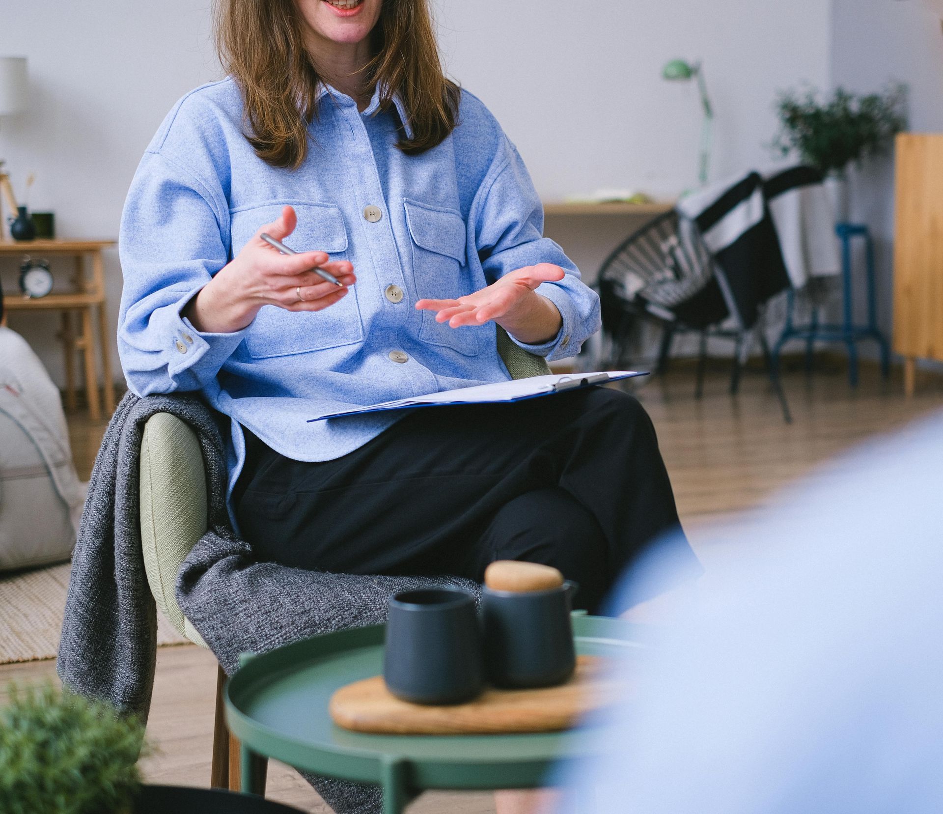 Woman in blue shirt gestures, holding clipboard, talking in a counseling session. Small table with dark mugs, neutral background.