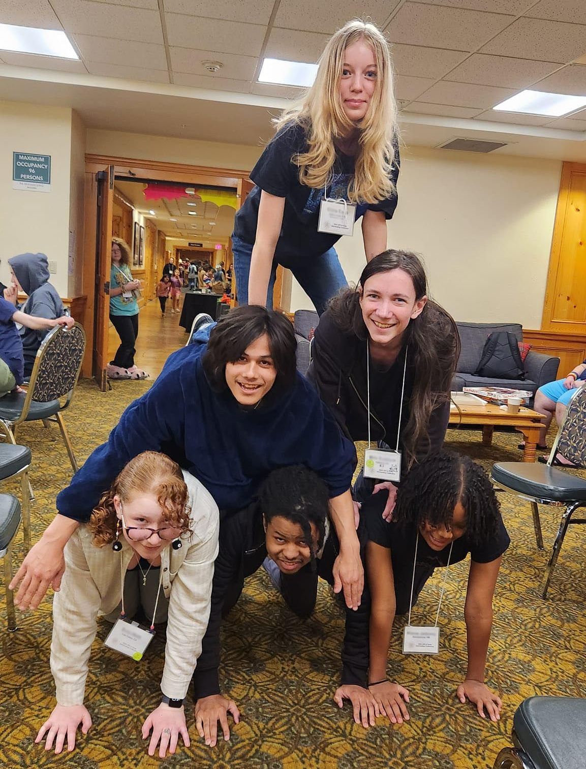 People forming a human pyramid indoors, smiling, with a hallway and chairs in background.