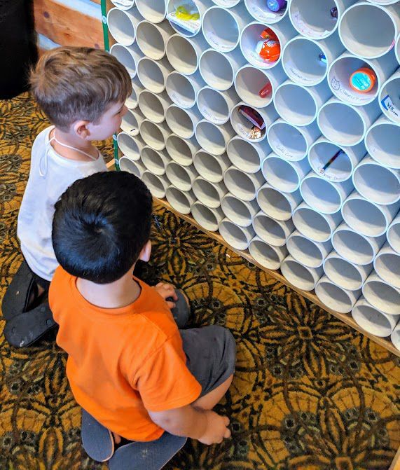 Two children looking at toys inside a white PVC pipe wall. One wears orange, the other white.