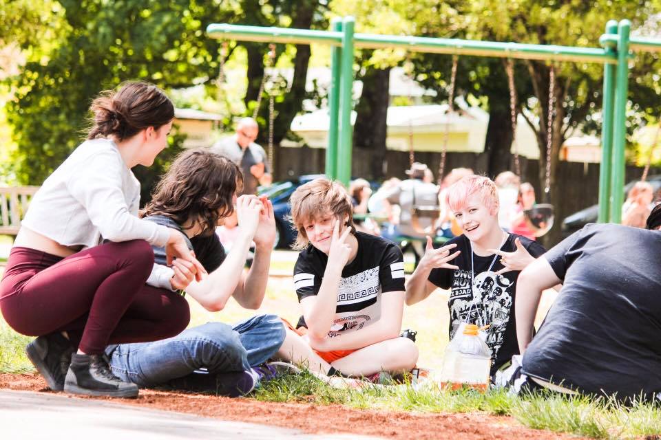 A group of young people are sitting on the ground in a park talking to each other.