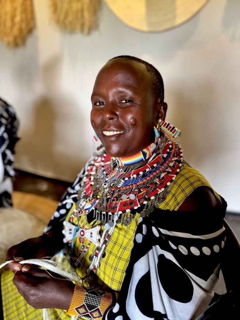 Woman smiles, wearing colorful beaded jewelry and patterned clothing, sitting indoors, weaving.
