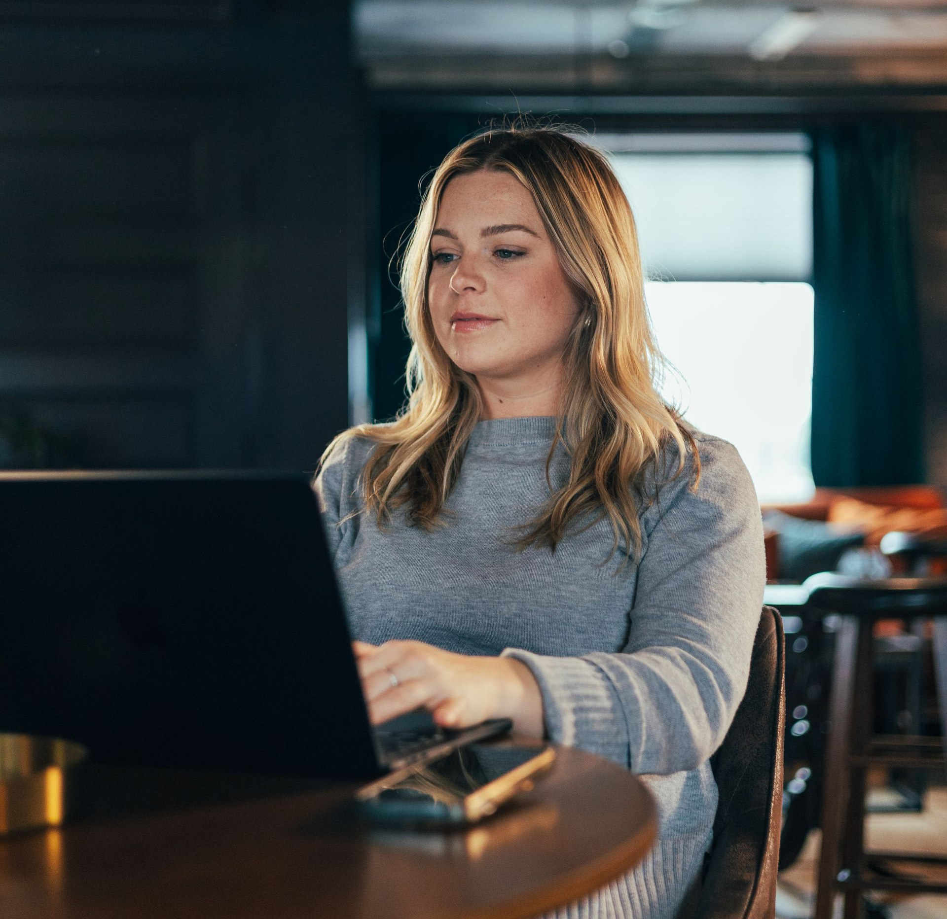 Woman seated at a round table, working on a laptop in a cafe setting.