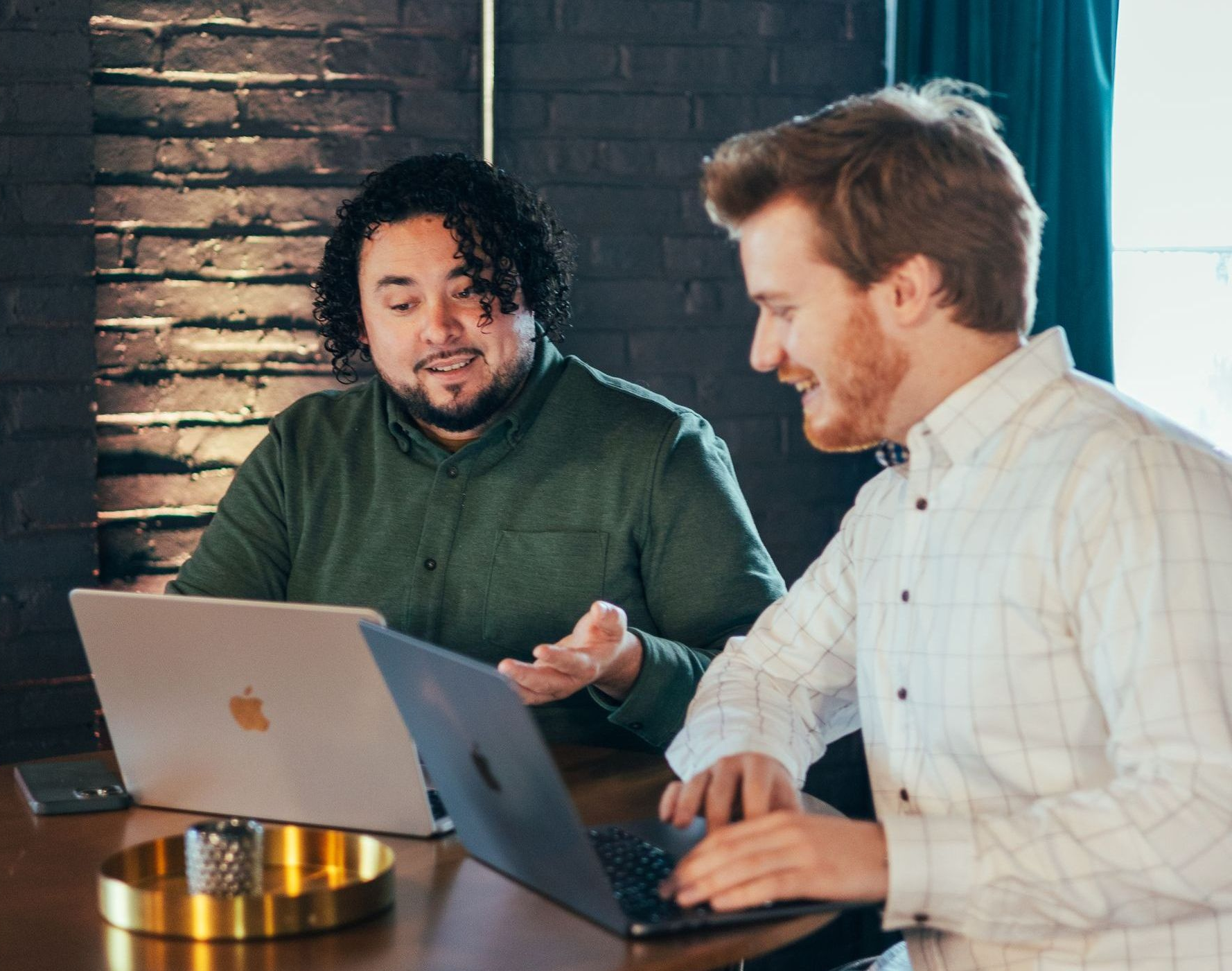 Two men at laptops, one gesturing, the other smiling, by a brick wall.