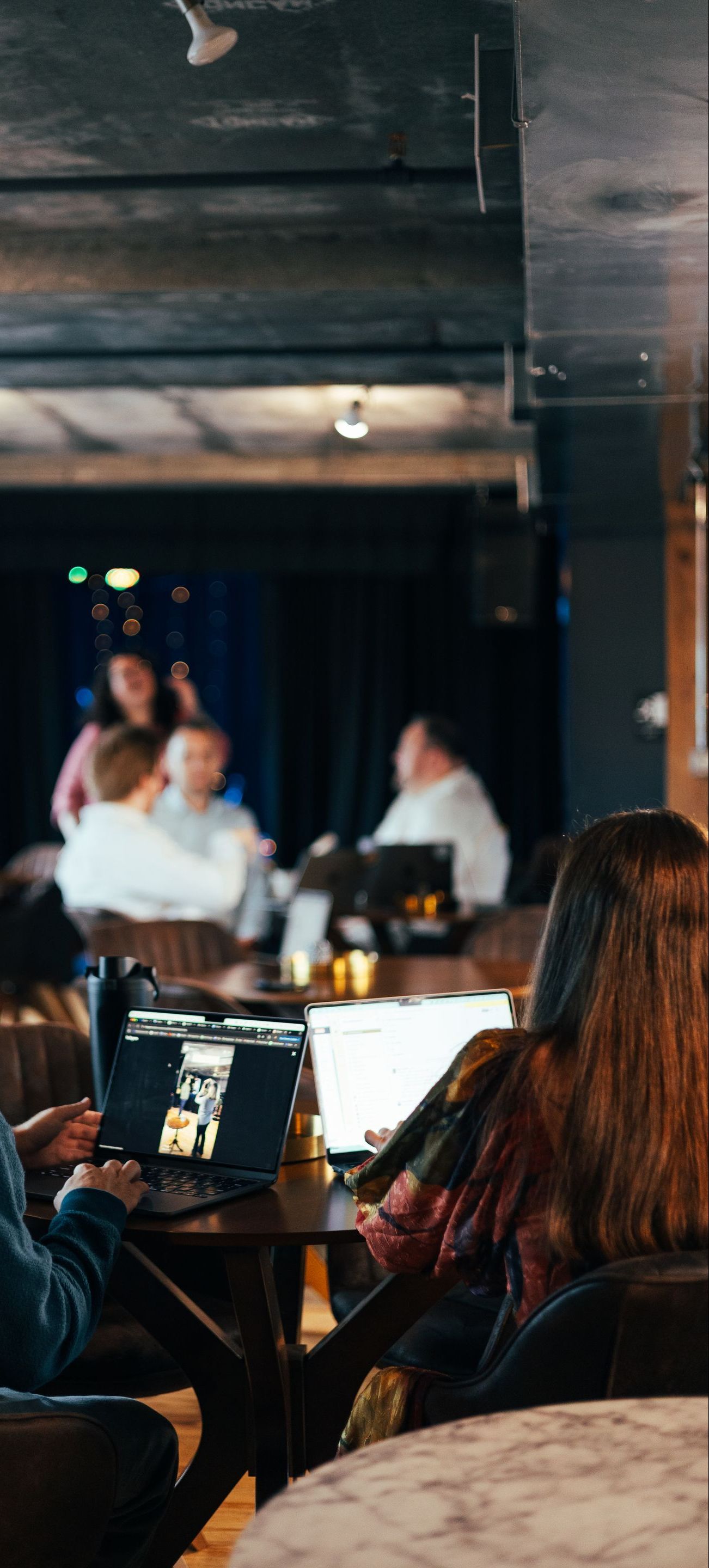 People working on laptops in a dimly lit cafe. Others are conversing in the background.