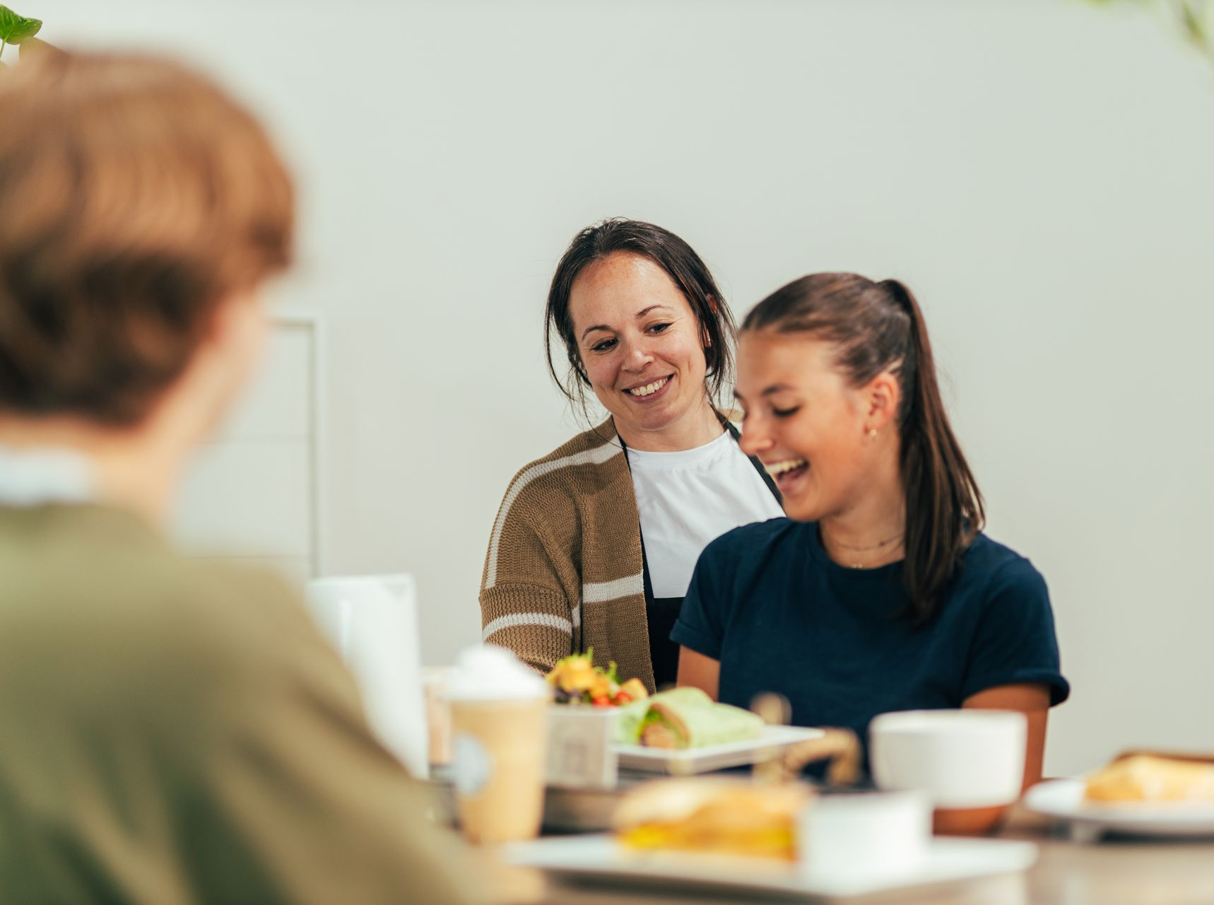 Two women and a person with their back to the camera, laughing and smiling at a table with food.
