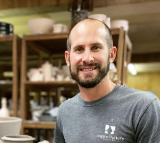A bald man with a beard wearing a grey shirt that says inspire pottery