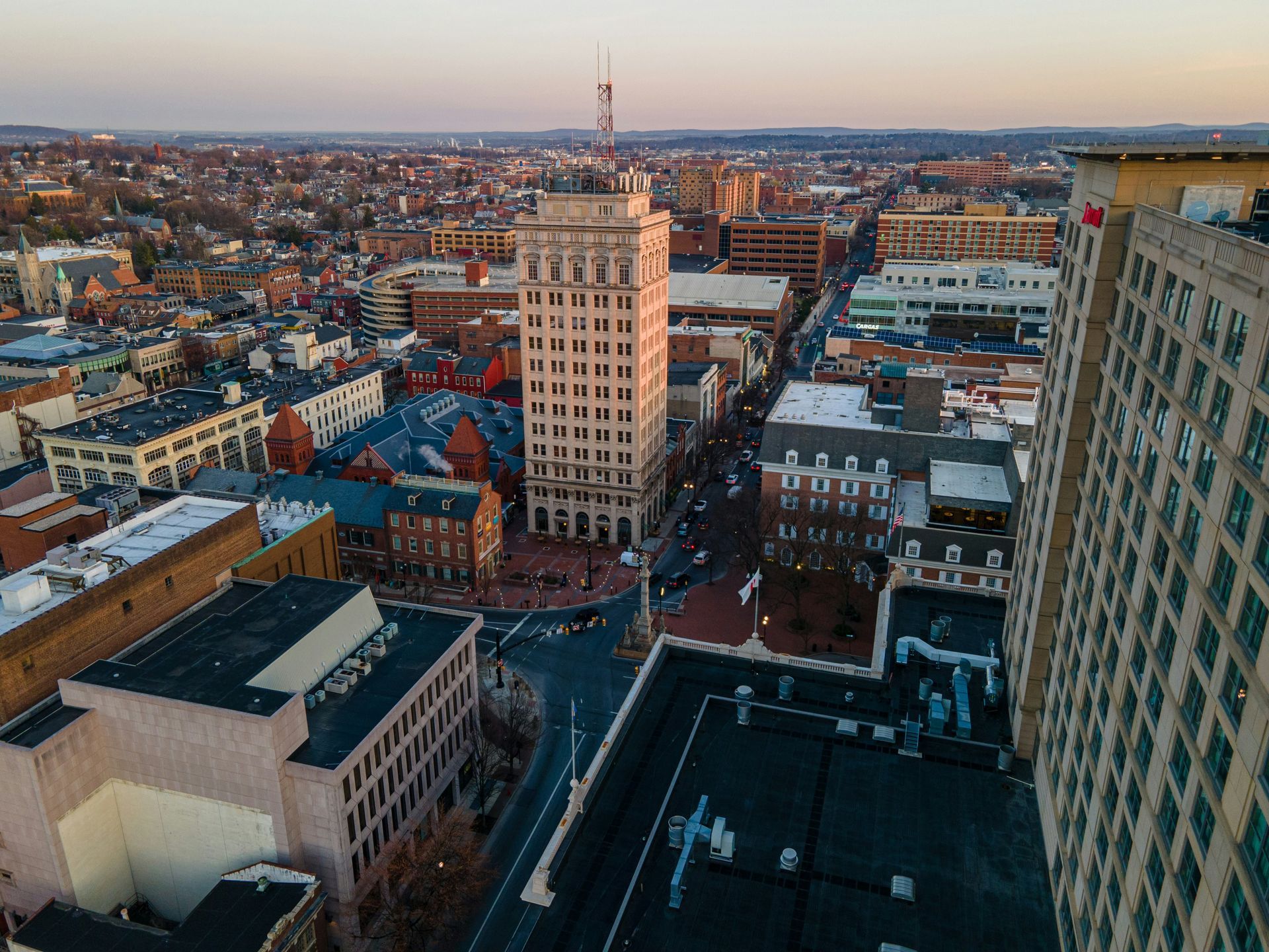 Aerial view of a city downtown at sunset; tall buildings line a main street; brown, tan, and red brick structures.