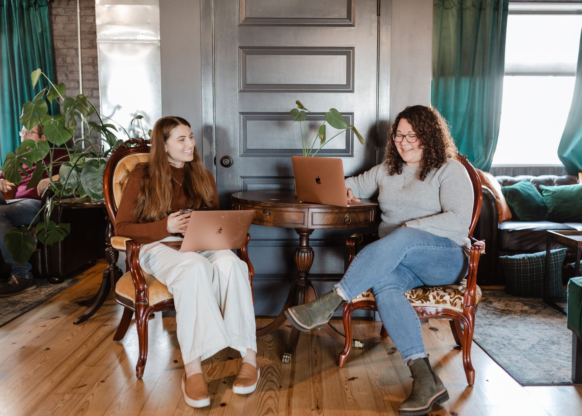 Two women are sitting in chairs with laptops in a living room.
