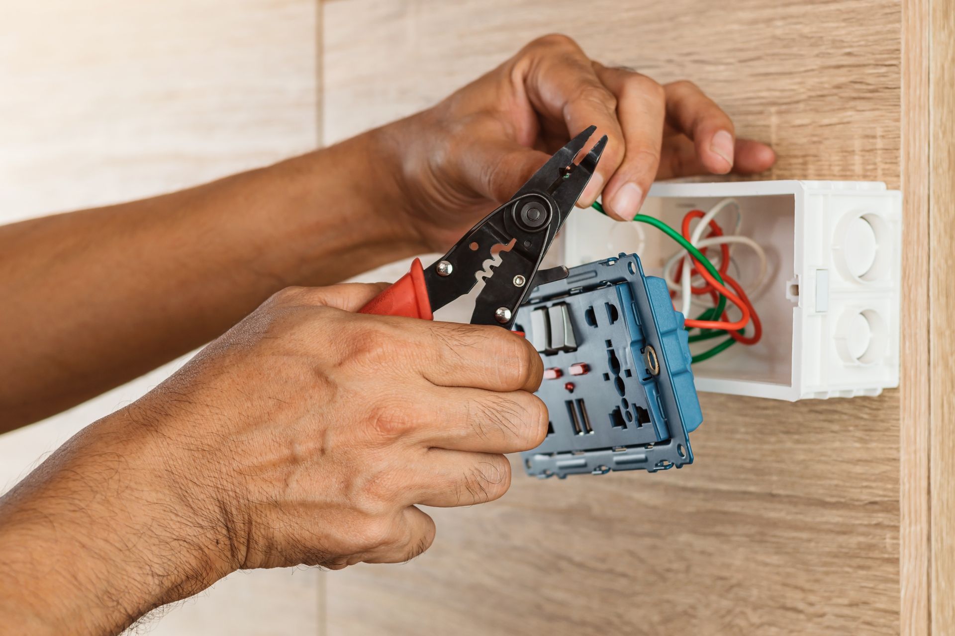 Hands wiring a wall electrical outlet with pliers and exposed red and green wires