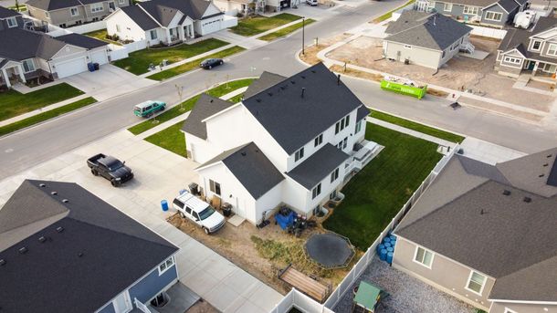 Aerial view of a white two-story house with a driveway, parked cars, and neighboring homes in a suburban street.