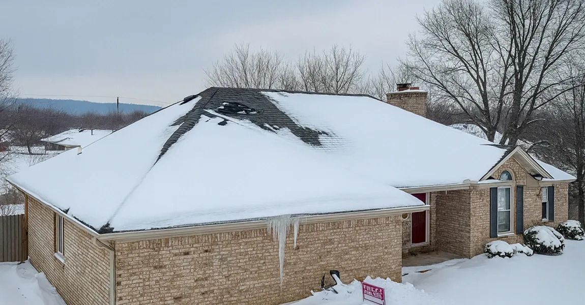 A brick house partially covered in snow and ice with large icicles hanging from the roofline on a winter day.