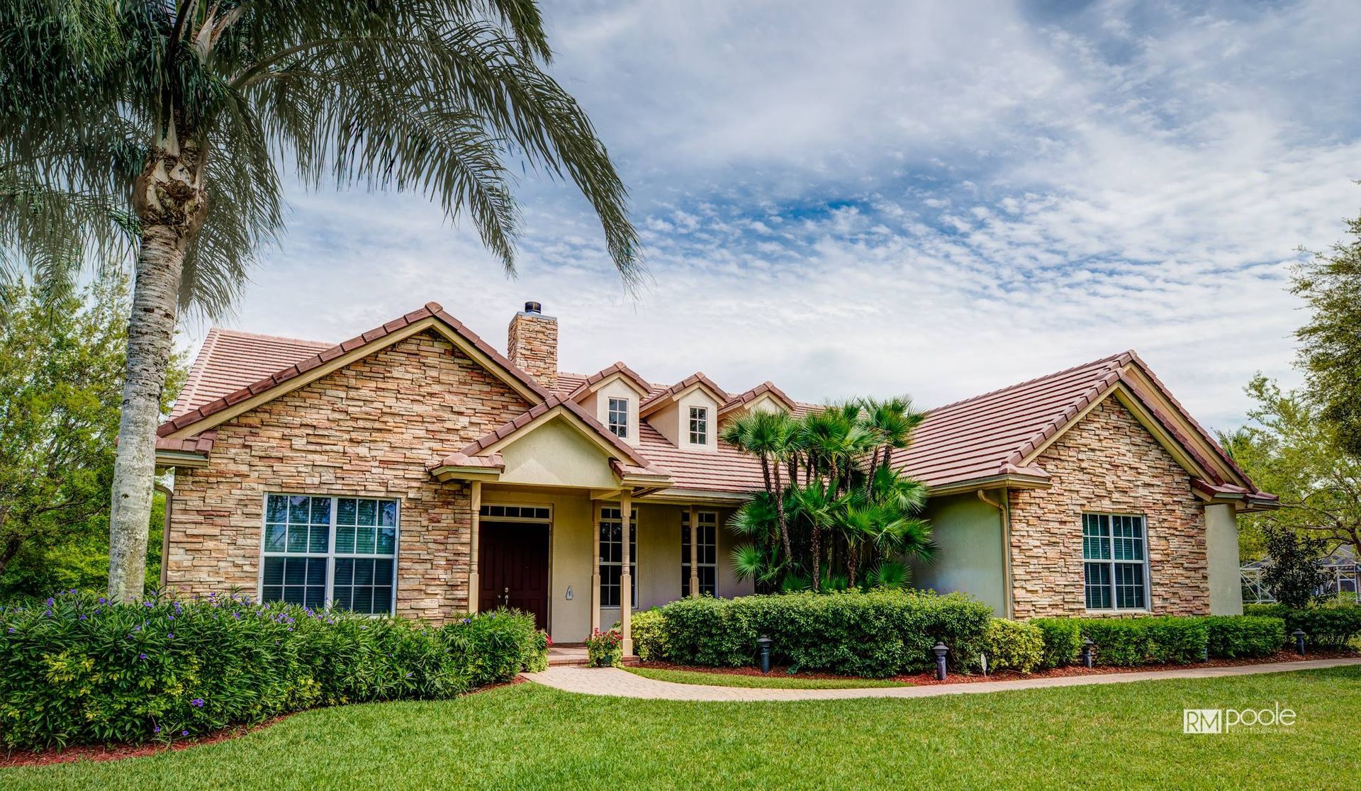 A tan stone house with a red tiled roof and brown front door, surrounded by green landscaping and a palm tree.