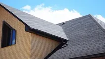 The upper portion of a tan brick house with a dark gabled roof, a triangular window, and black gutters against a blue sky.