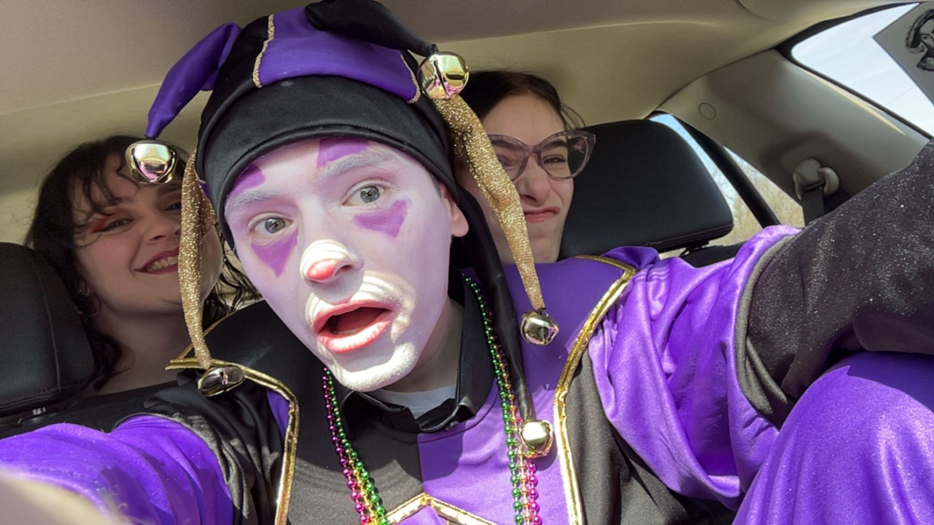 Three people in purple costumes pose inside a car, with one person taking a selfie and two wearing face paint.