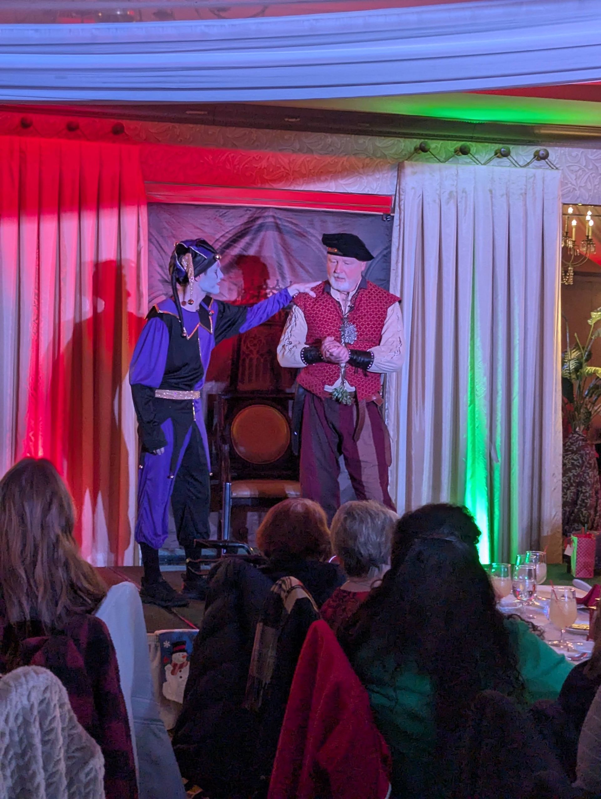 Two performers on a lit stage, one in a black hat and red vest, speaking to a microphone before an audience.