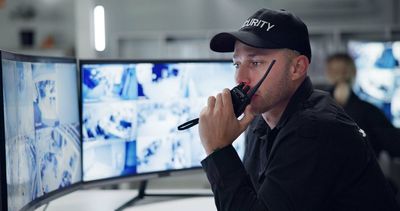 A security guard is talking on a walkie talkie while looking at a monitor.