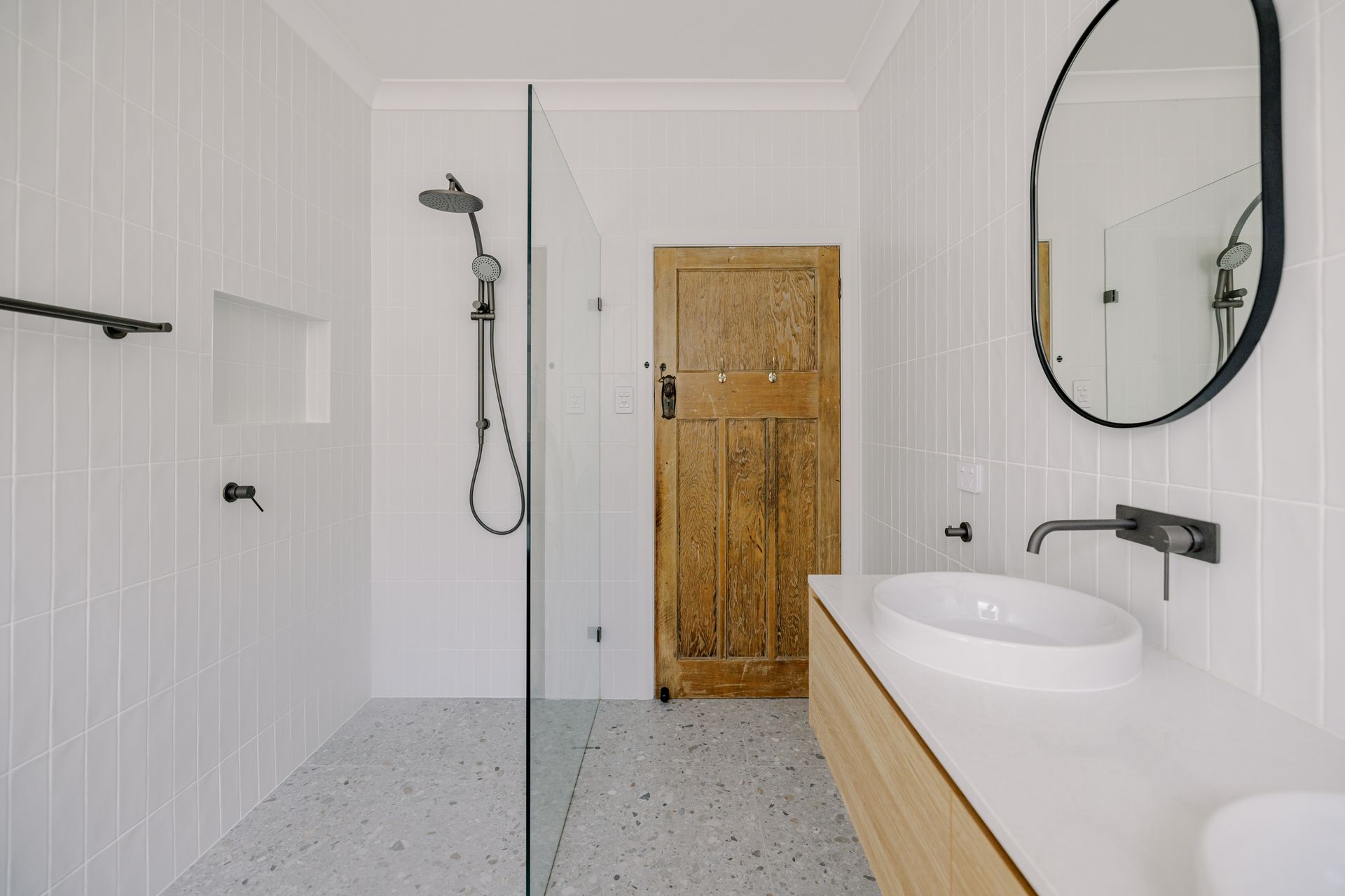 White bathroom with wood door, glass shower, oval mirror, and vanity.