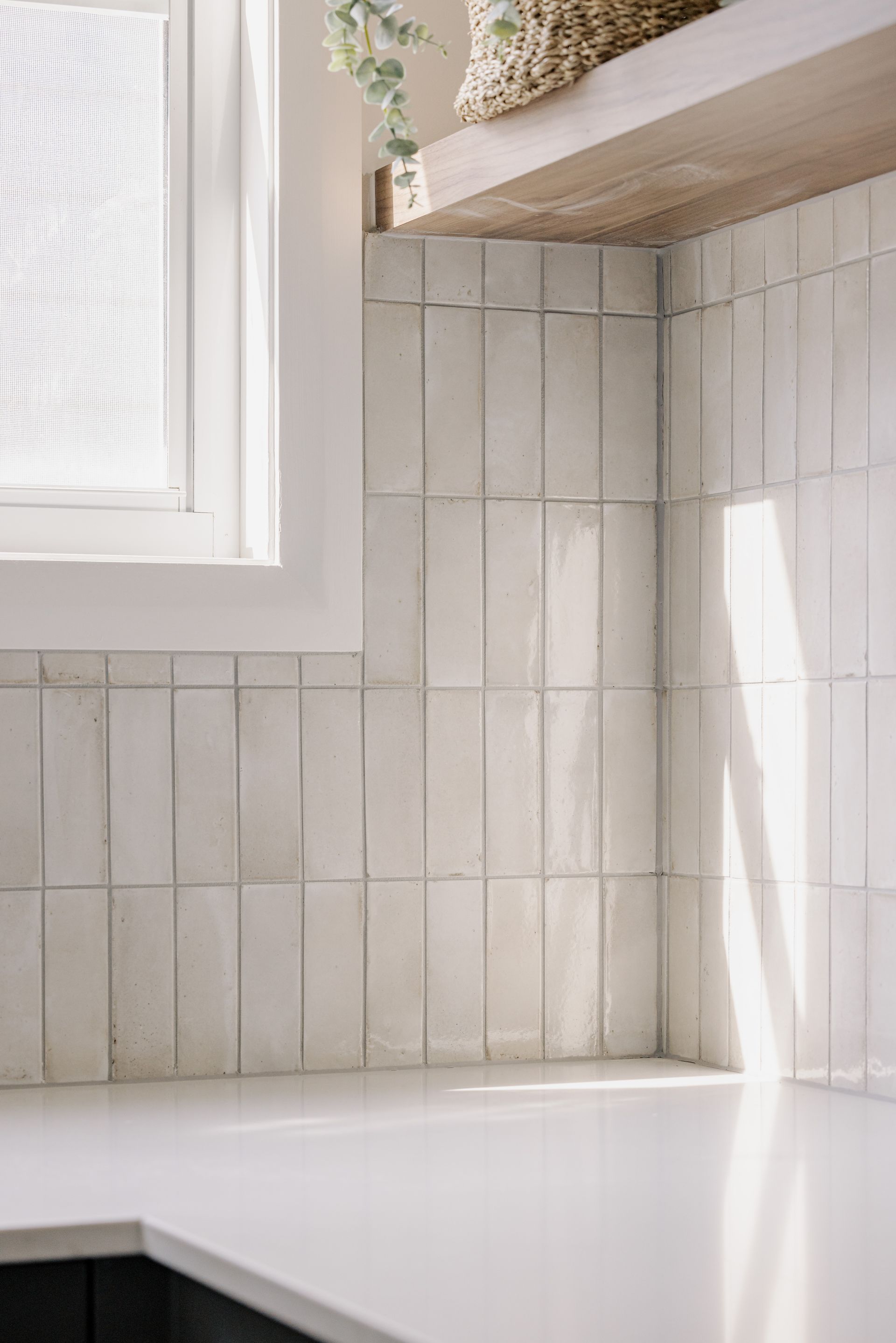 White tiled kitchen backsplash with a white countertop under a window and a wooden shelf.