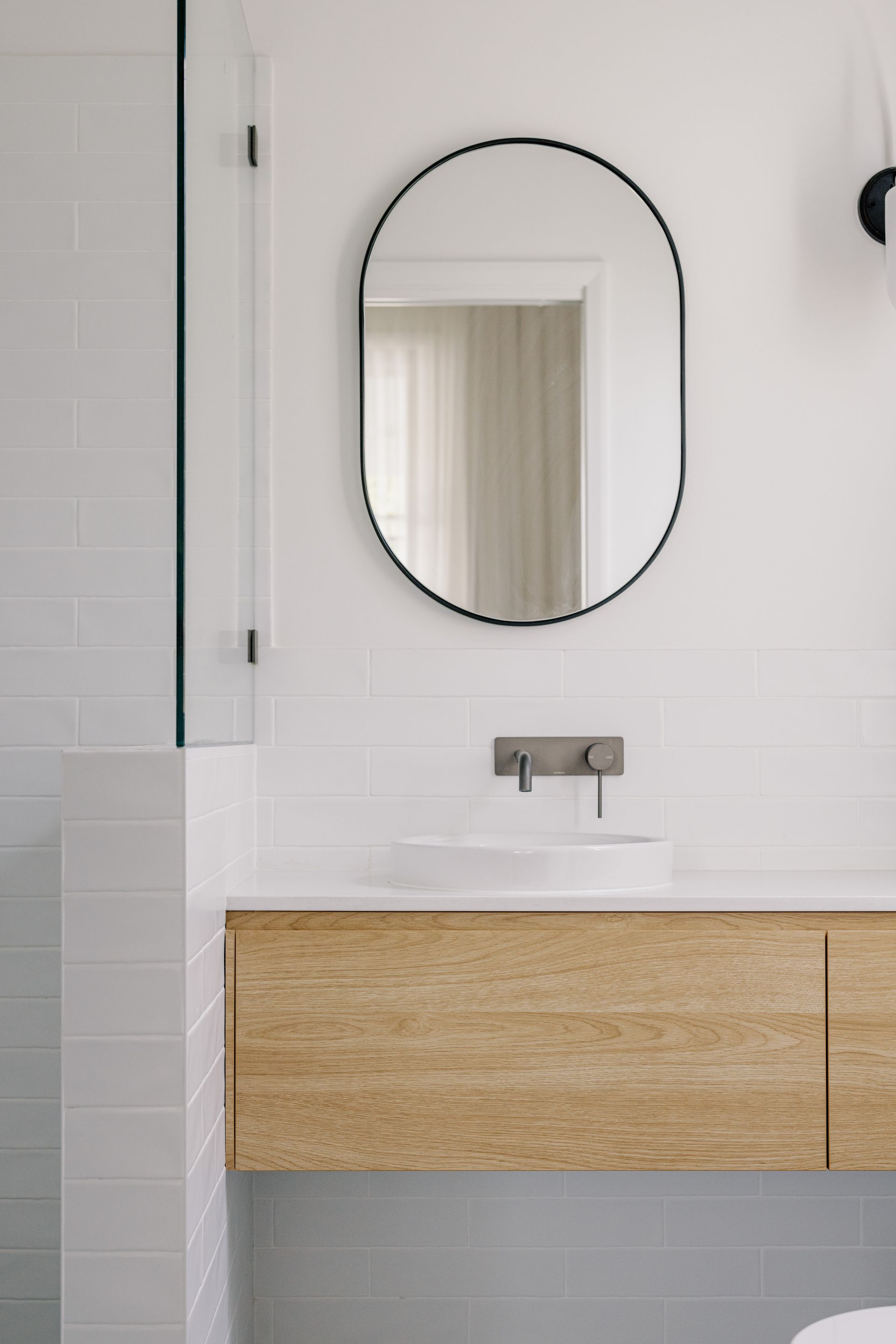 Modern bathroom with wooden vanity, white sink, oval mirror, and white tile.