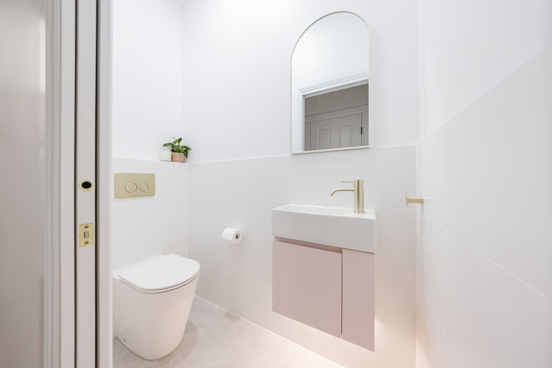 Small, modern white powder room with pink cabinet, gold fixtures, and arched mirror.