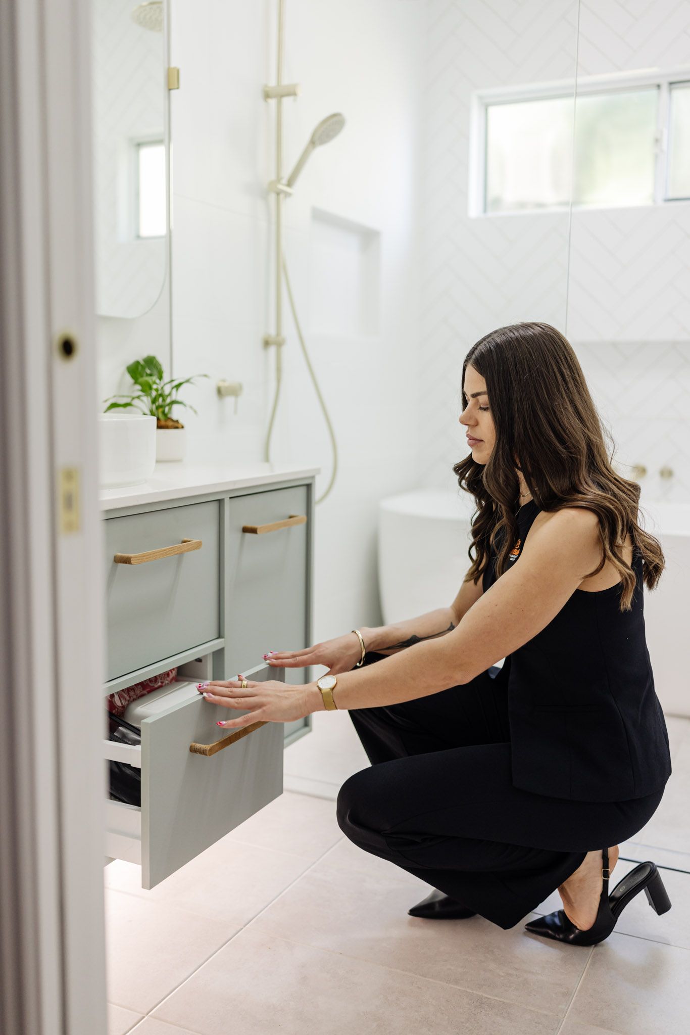 Woman opening a bathroom vanity drawer, dressed in black. Light gray vanity, gold hardware, white walls, light pink floor.