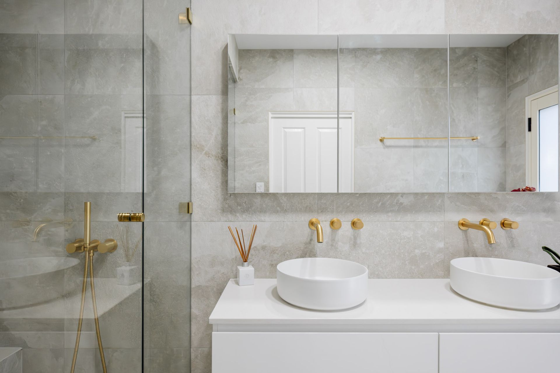 Modern bathroom with gold fixtures, dual sinks, large mirror, and glass shower.
