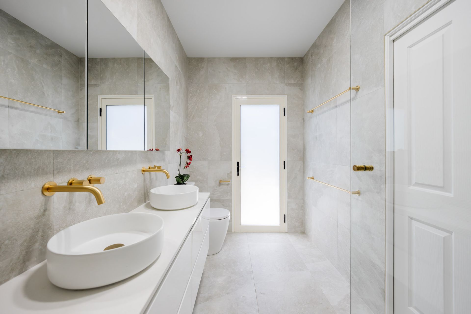 Modern white bathroom with gold fixtures, dual sinks, and a frosted glass door.