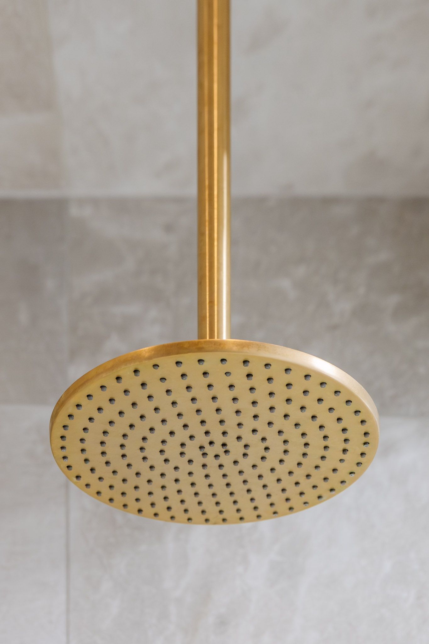Gold-colored rain shower head mounted on a vertical pipe, with many small holes, against a tiled wall.
