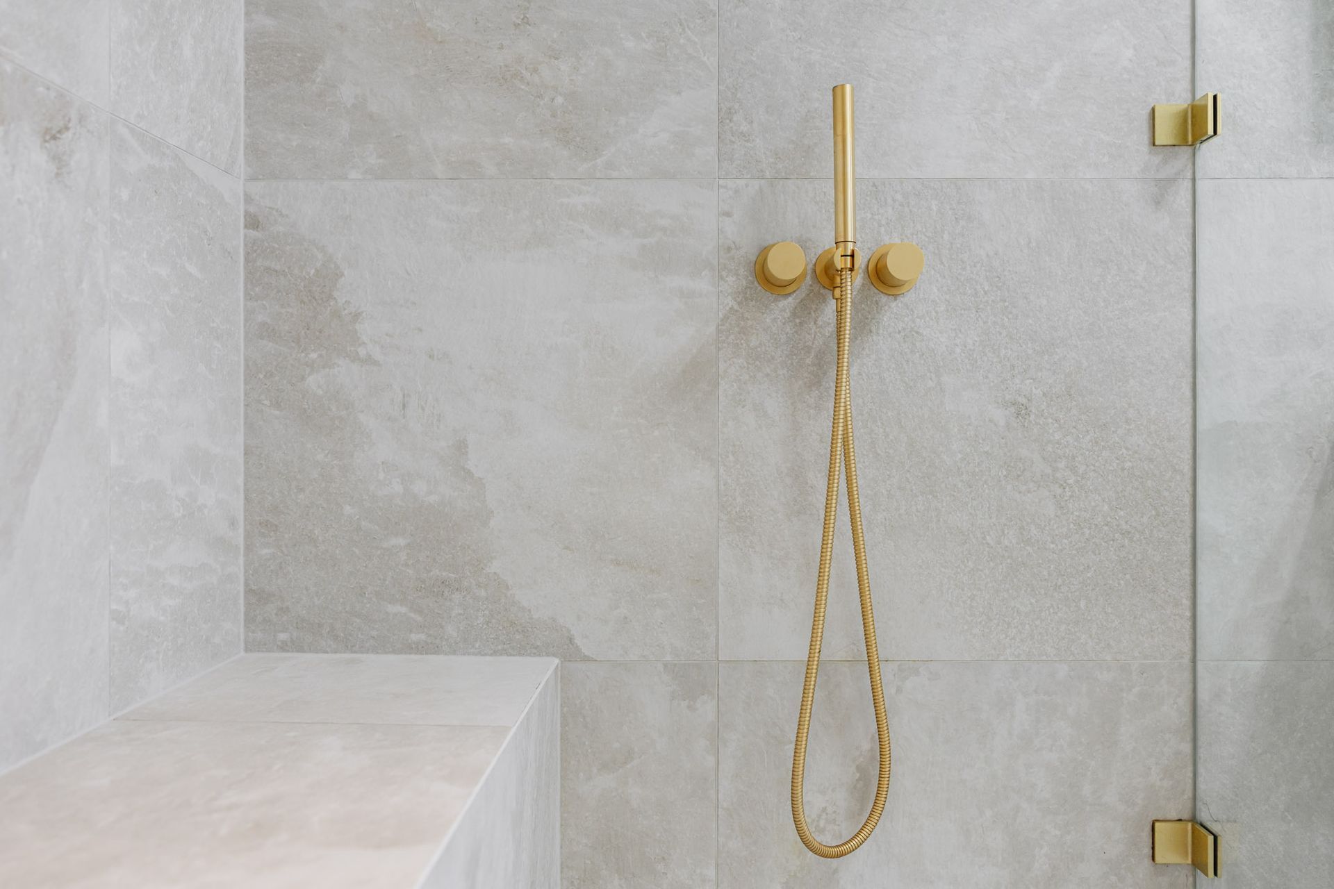 Gold showerhead and fixtures on a tiled wall, near a glass shower door and bench.