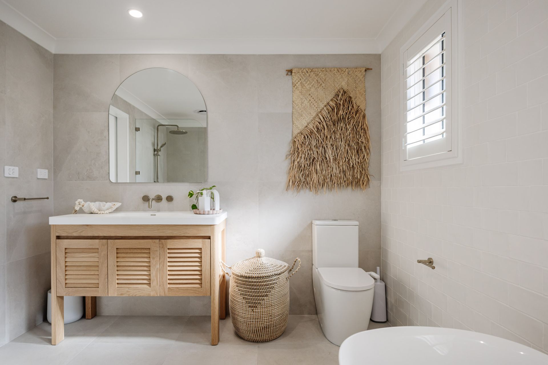Bathroom with wooden vanity, arched mirror, neutral walls, woven wall hanging, and toilet.