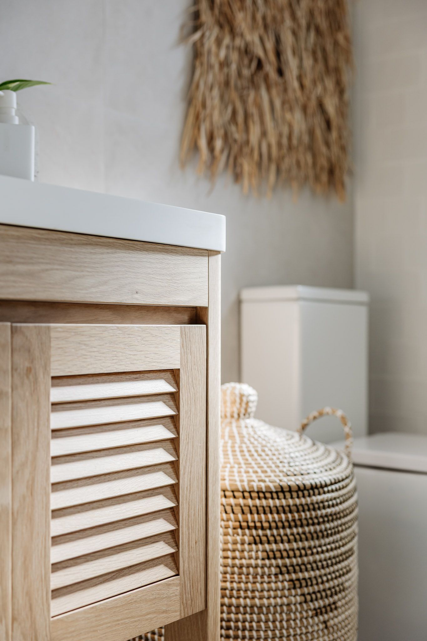 Light wood vanity cabinet with louvered door, woven basket, and toilet in a neutral-toned bathroom.