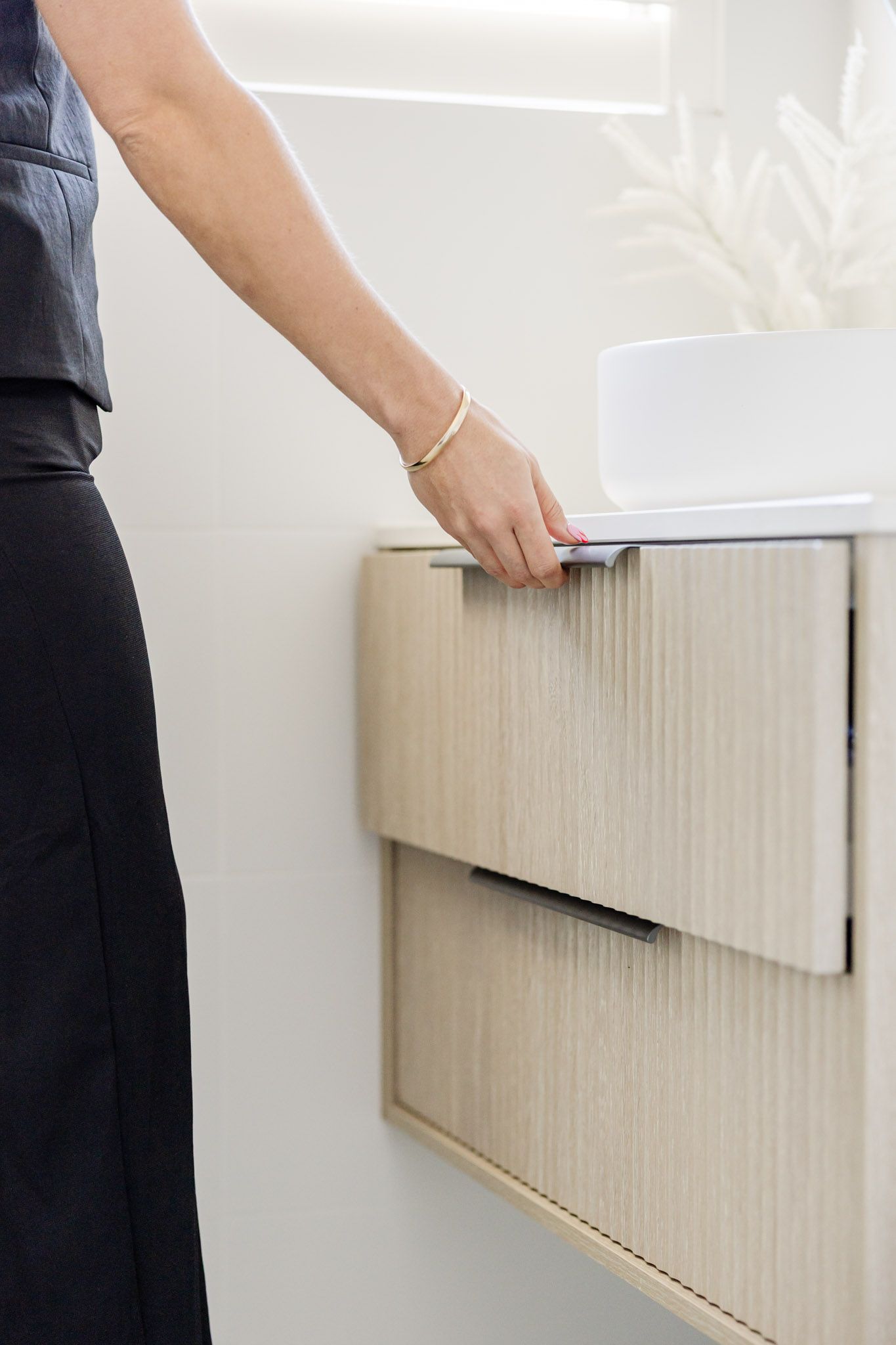 Person opening bathroom drawer. Light wood cabinet, white wall, porcelain sink.