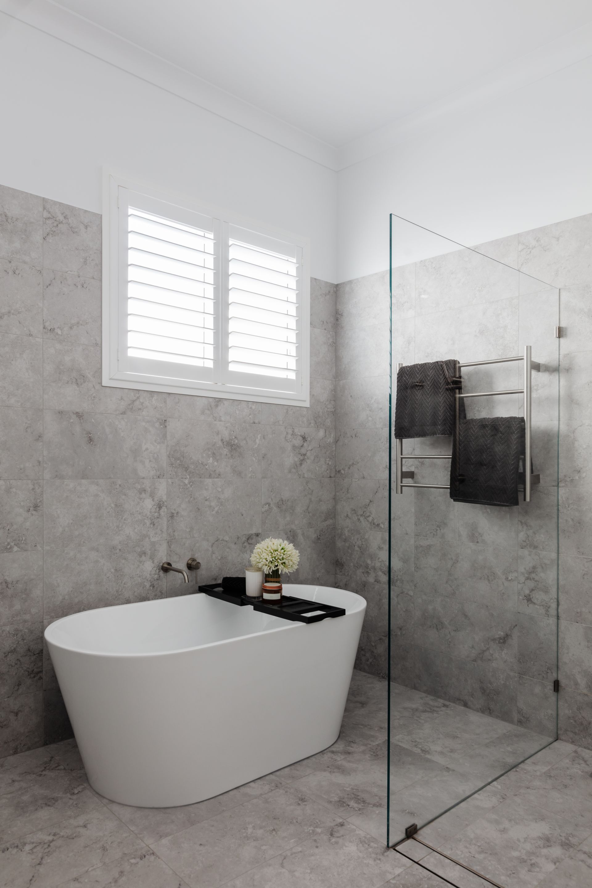 Modern bathroom with gray tiled walls, white tub, glass shower, and white shutters.