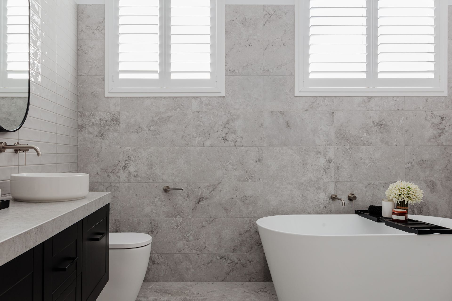 Bathroom with gray textured wall tiles, white bathtub, and black vanity. Two windows with white shutters.