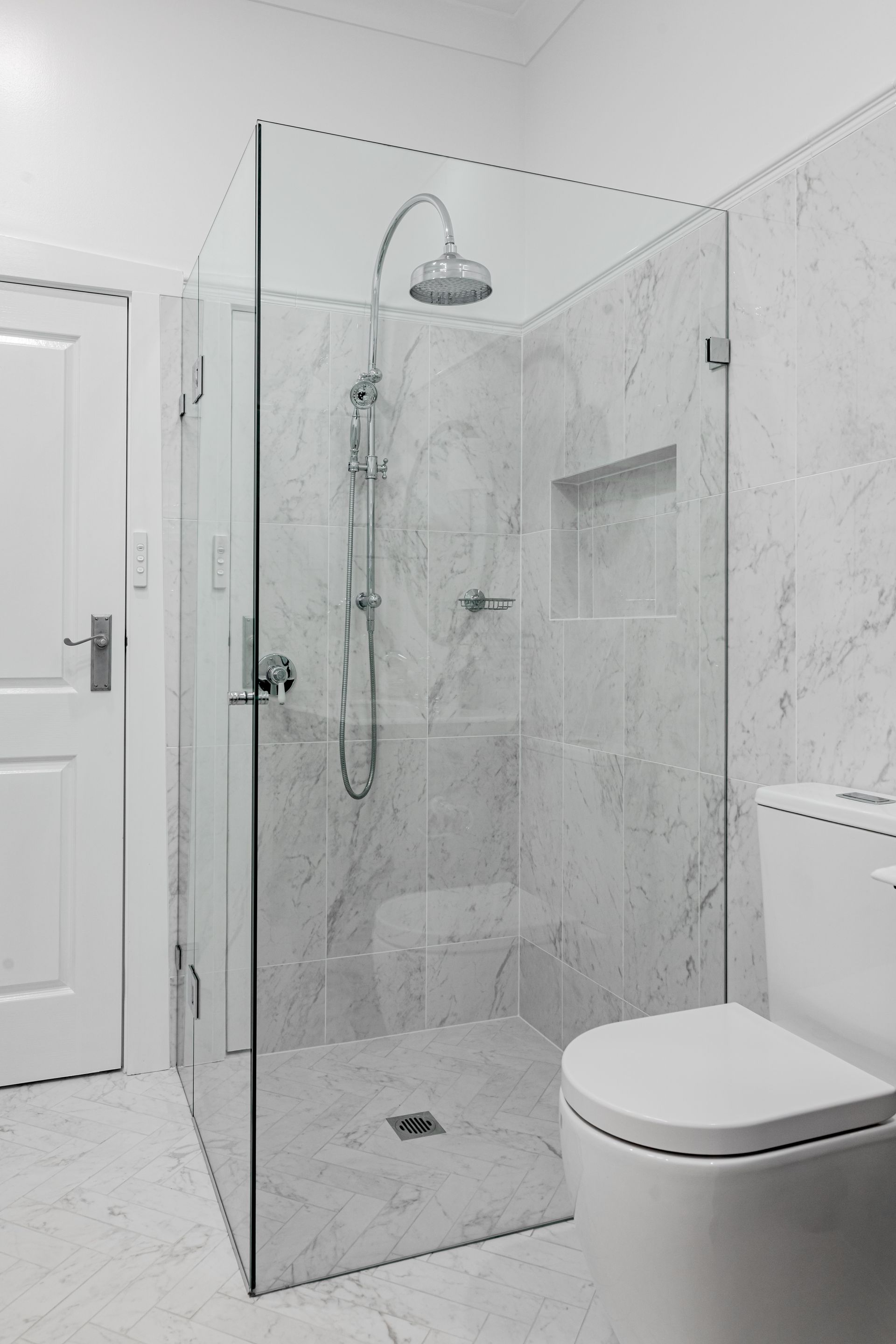 Bathroom with glass shower, white marble walls, toilet, and door.