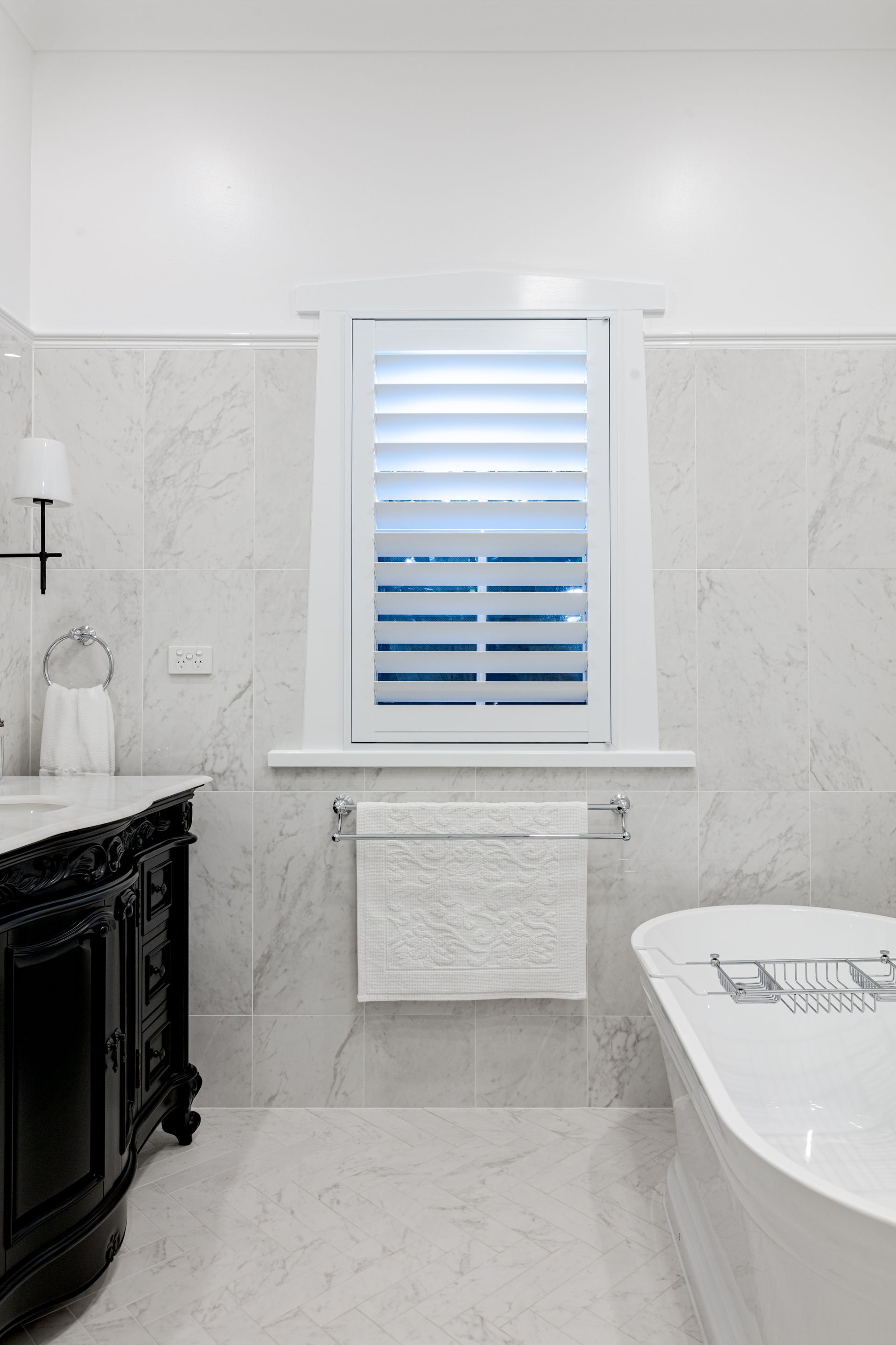 Bathroom with marble tiles, white shutters, black vanity, and a bathtub.