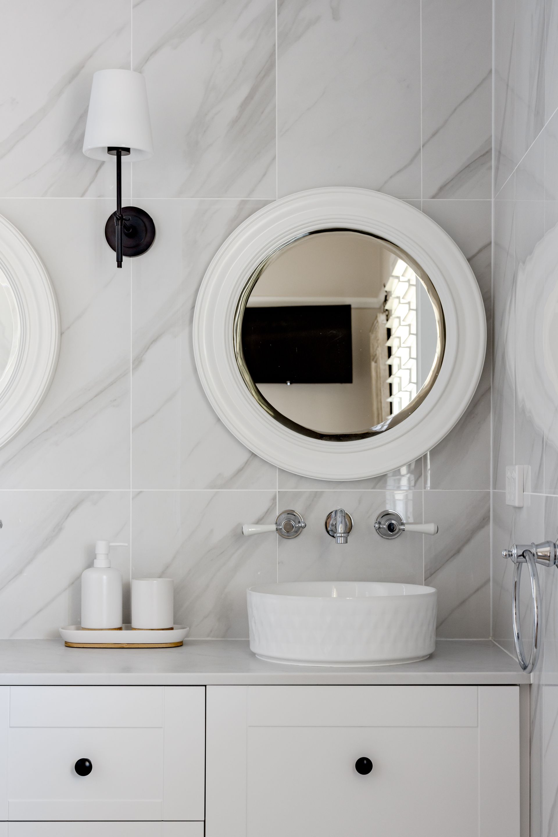 White bathroom with a round mirror over a sink, featuring marble tile and black accents.