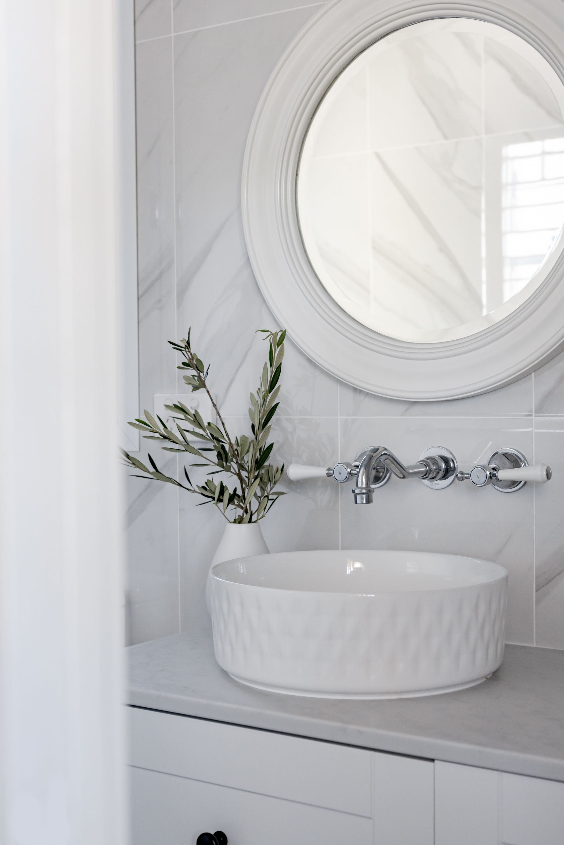 White bathroom with a decorative sink, round mirror, and vase of greenery.