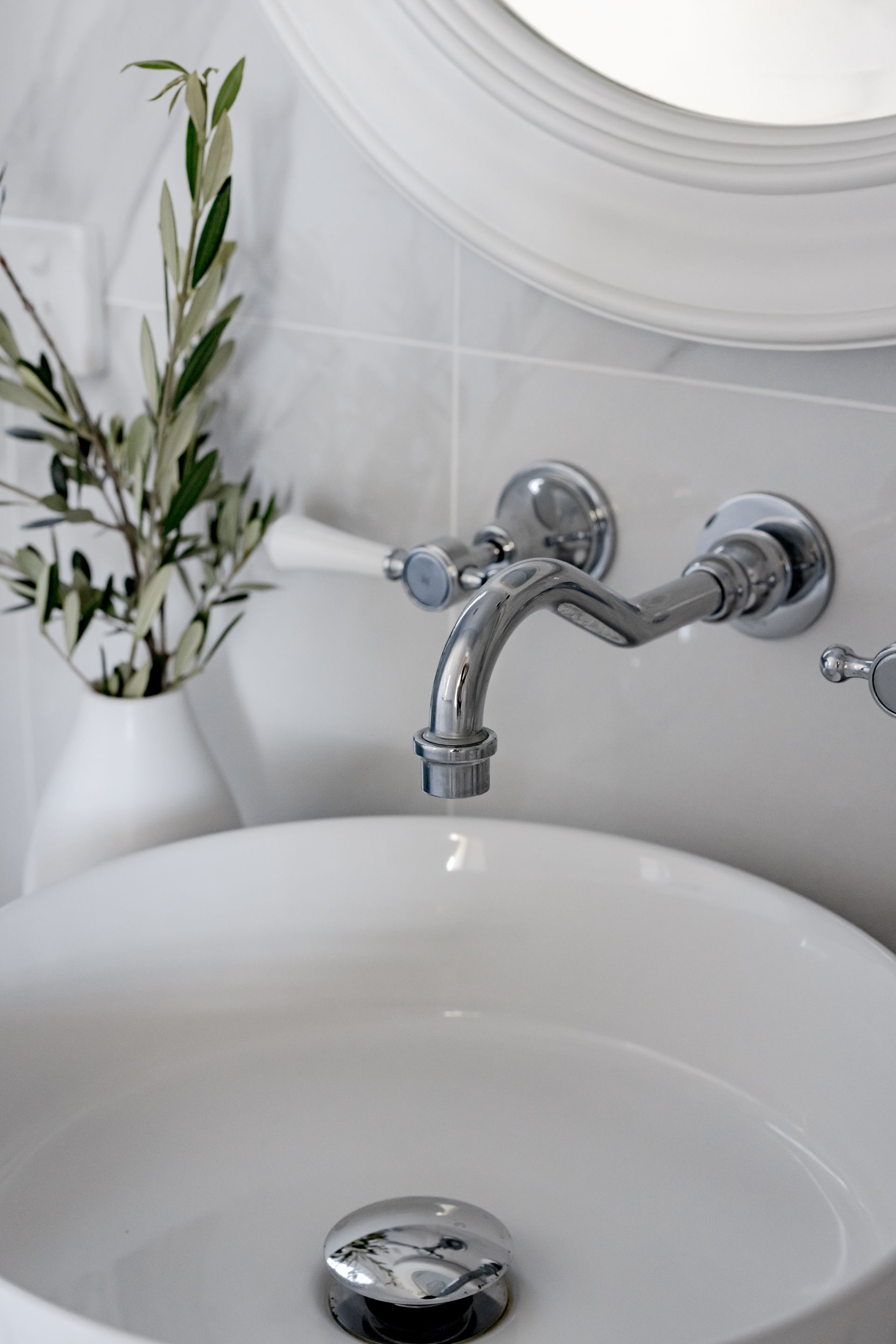 White sink with chrome faucet and olive branch in vase against a gray tiled wall.