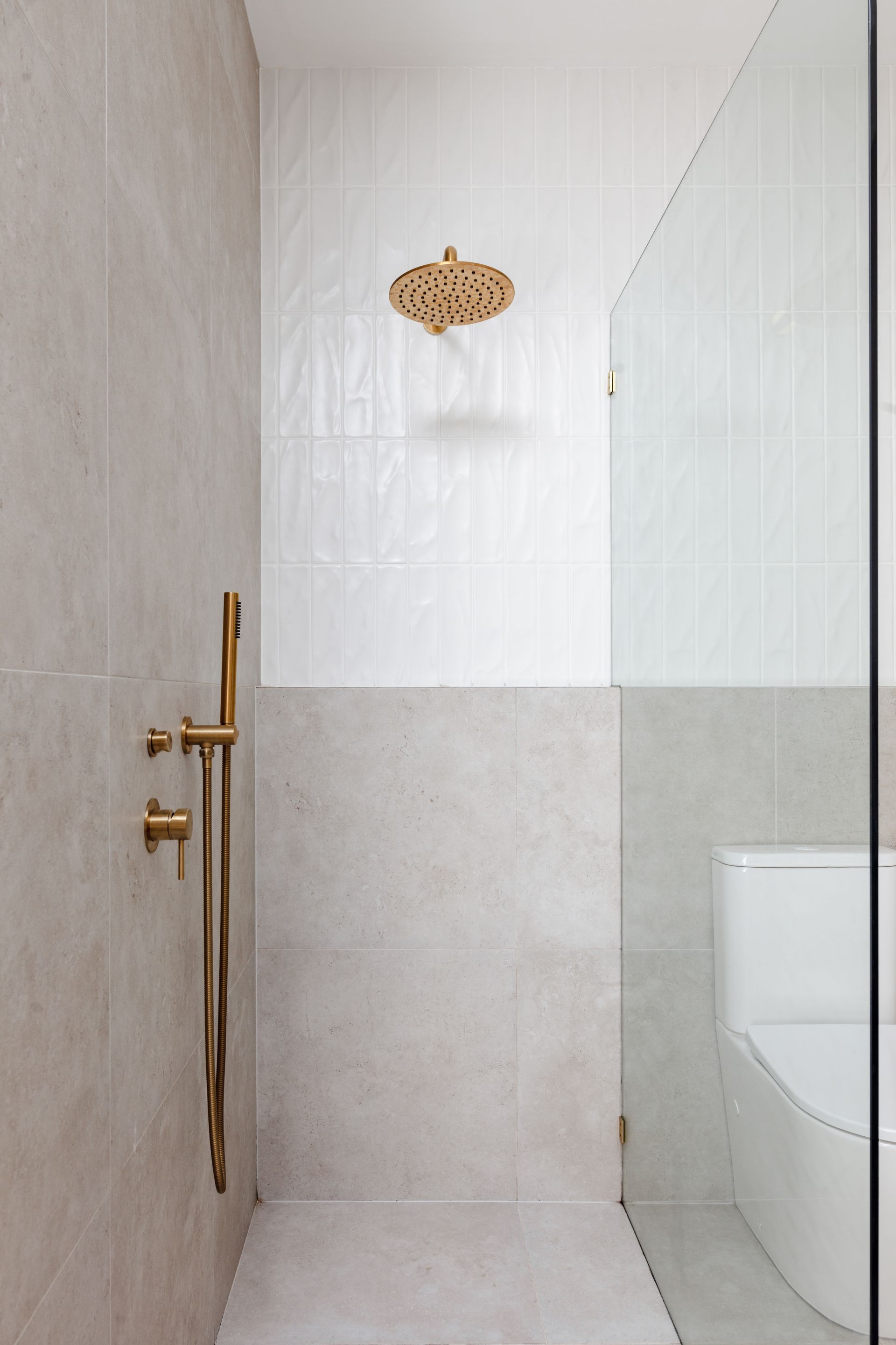 Shower with gold fixtures and white and beige tile, next to a toilet behind glass.