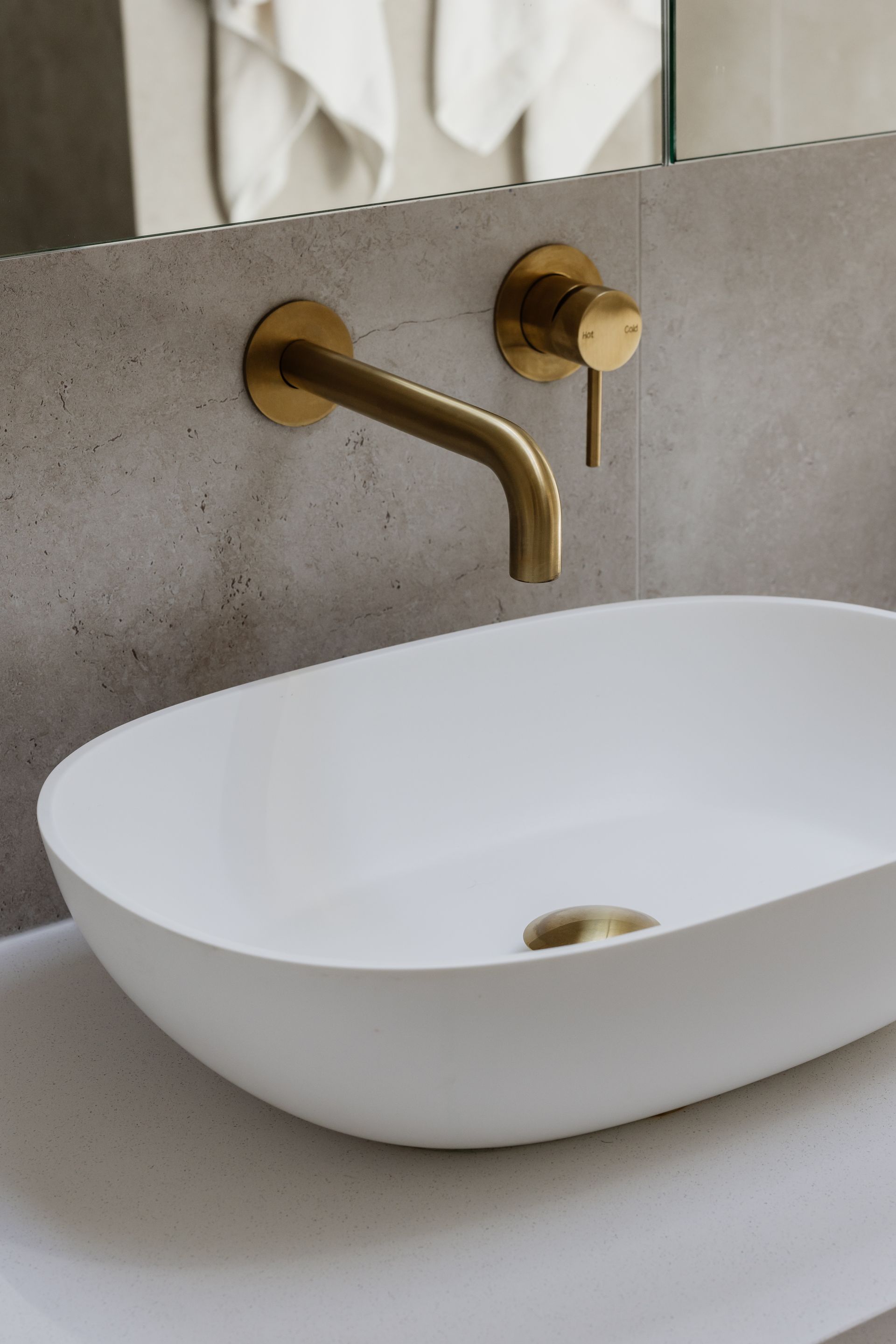White oval sink with gold faucet against a gray textured wall.