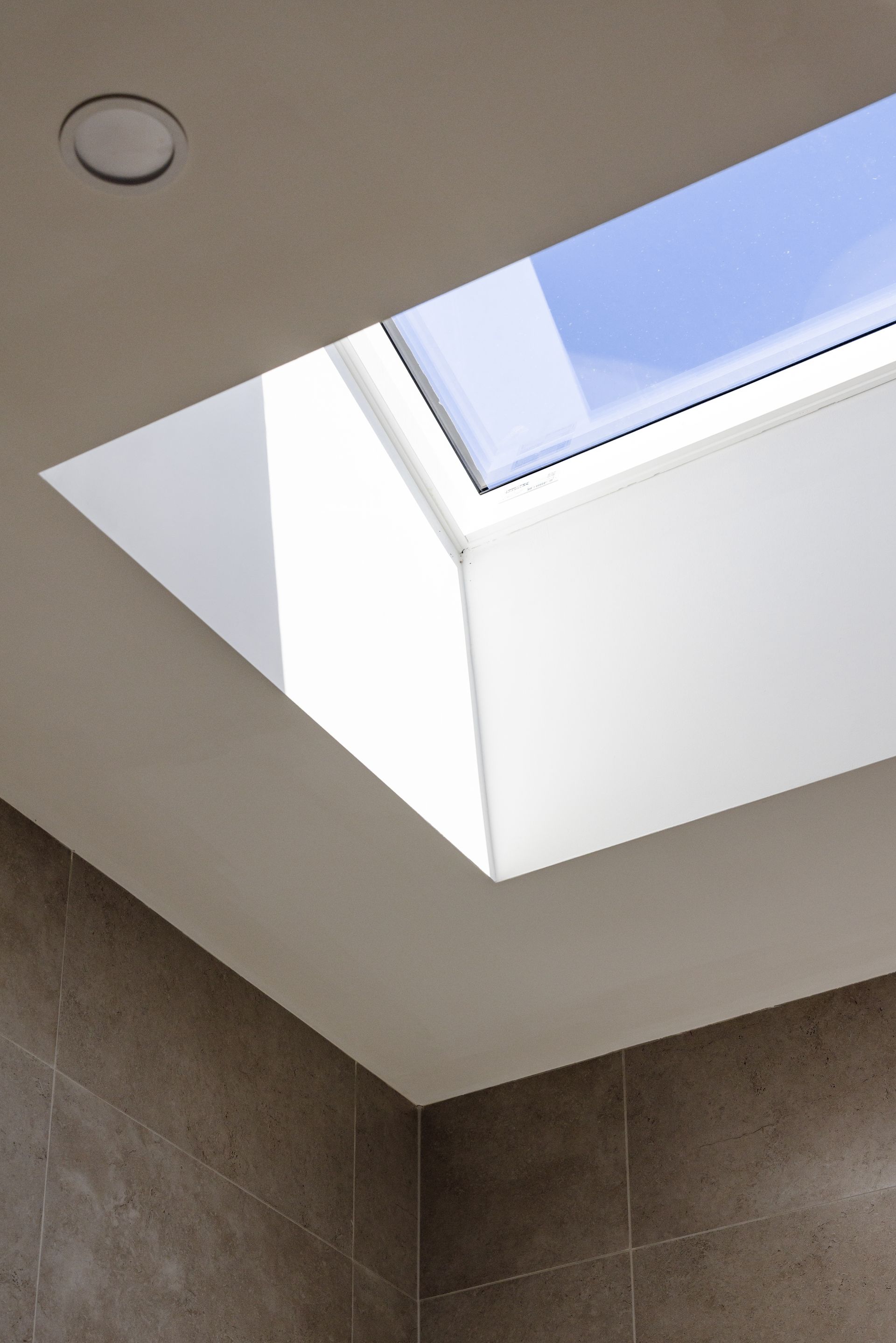 Skylight in a white-walled corner of a room, with a view of blue sky and a small recessed light.
