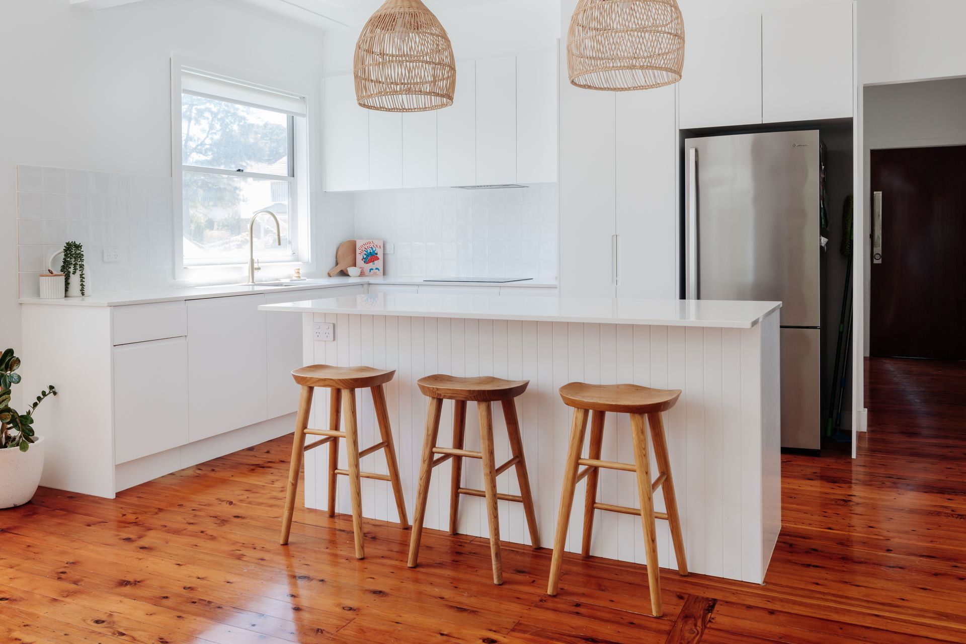 White kitchen with wood stools, island, and pendant lights, on a wooden floor.