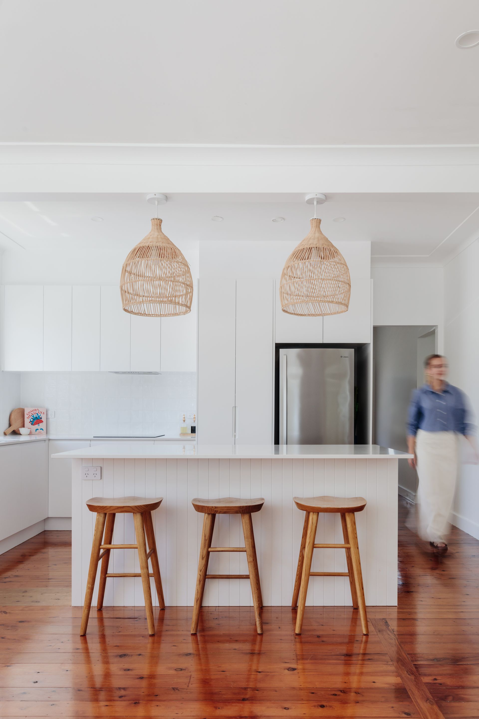 White kitchen with island, three wooden stools, woven pendant lights, person walking.