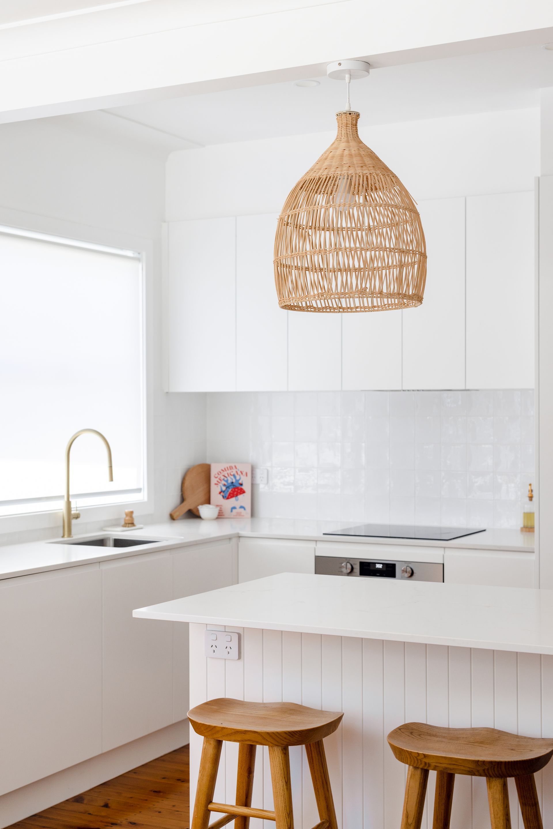 White kitchen with woven pendant light, white cabinets, island with wood stools, and gold faucet.