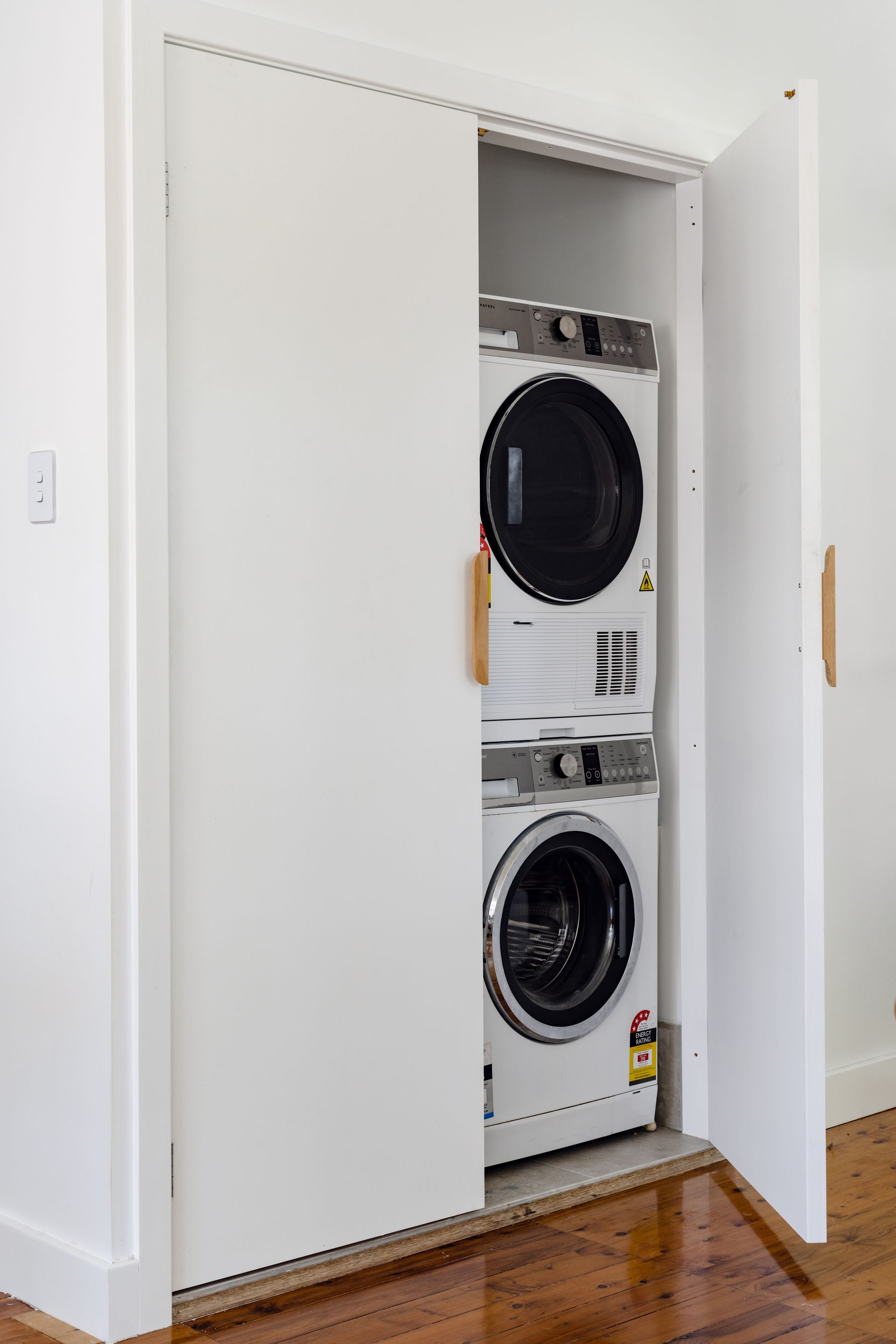 White, stacked washer and dryer in a closet with open doors and wooden handles.