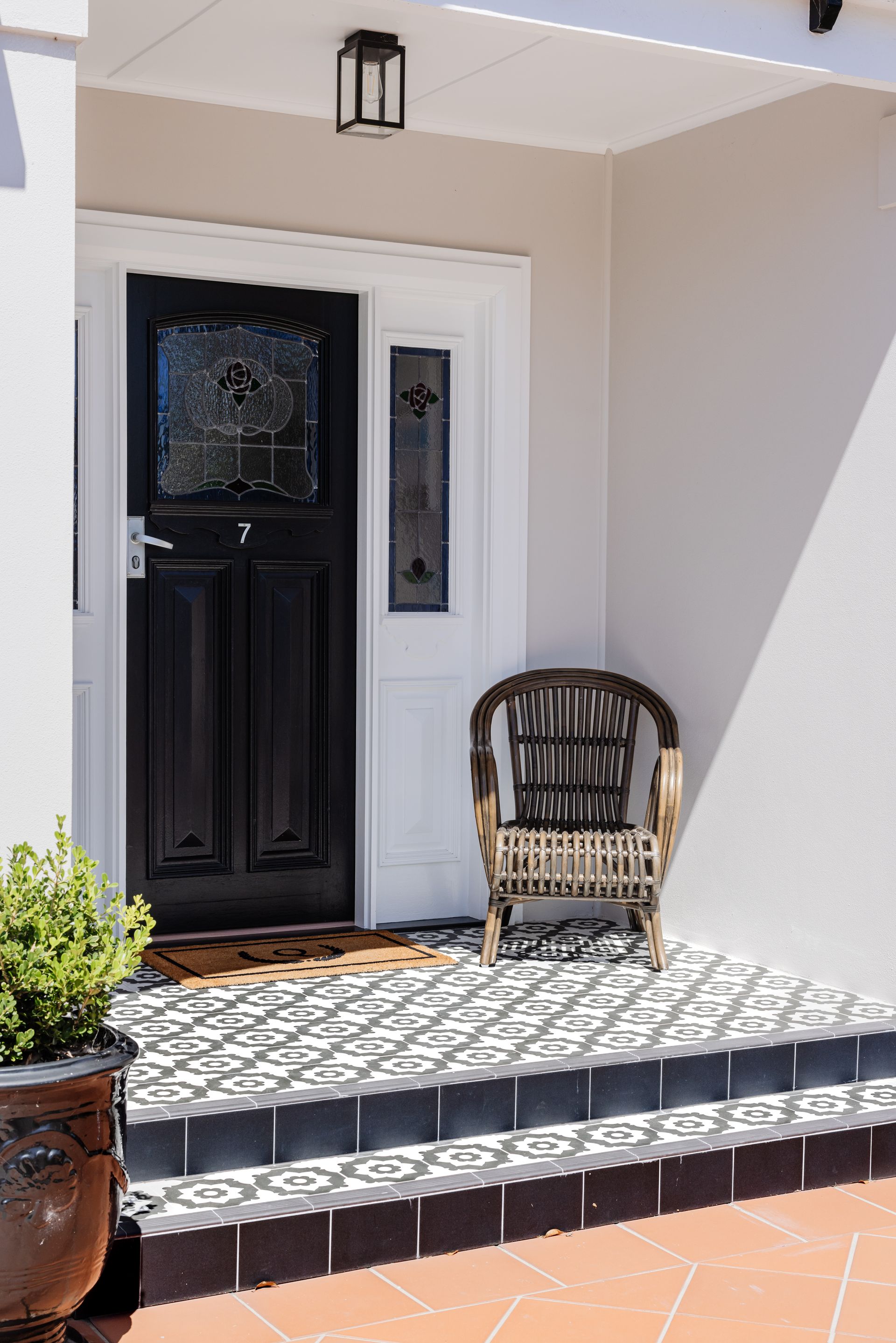 Black front door with patterned tile porch, wicker chair, and a potted plant.